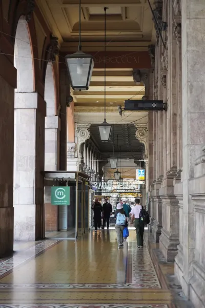 Una lunga passerella coperta con colonne ad arco conduce al Palazzo della Borsa. Il pavimento è decorato con colorate piastrelle a mosaico, e la luce del sole entra, proiettando ombre. Diverse persone si allontanano dall'osservatore lungo il portico.