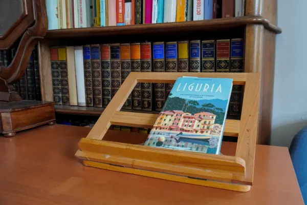 Un libro intitolato 'Liguria' è presentato su un cavalletto di legno, posizionato su una scrivania davanti a una grande libreria di libri antichi. La copertina del libro mostra una vista panoramica dell'architettura ligure e una barca.