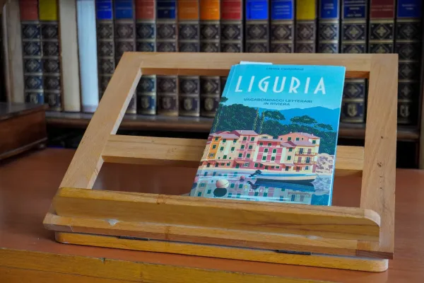 Un libro con un'illustrazione colorata di case costiere liguri e una barca su un supporto in legno, con una libreria di libri antichi sullo sfondo.