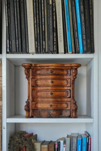 Una vista ravvicinata di una piccola cassettiera antica in legno con cinque cassetti, ognuno con una piccola manopola rotonda, appoggiata su una mensola bianca. Sopra la cassettiera, è visibile una fila di custodie per CD con titoli di musica classica. Sotto la cassettiera, un'altra mensola contiene una varietà di libri e un pezzo di metallo decorativo.