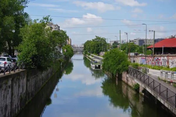 Un large canal à Milan, Italie, bordé d'arbres verts, de murs en pierre et de bâtiments modernes en arrière-plan. Le ciel et les arbres se reflètent dans l'eau calme, et un pont enjambe le canal au loin.