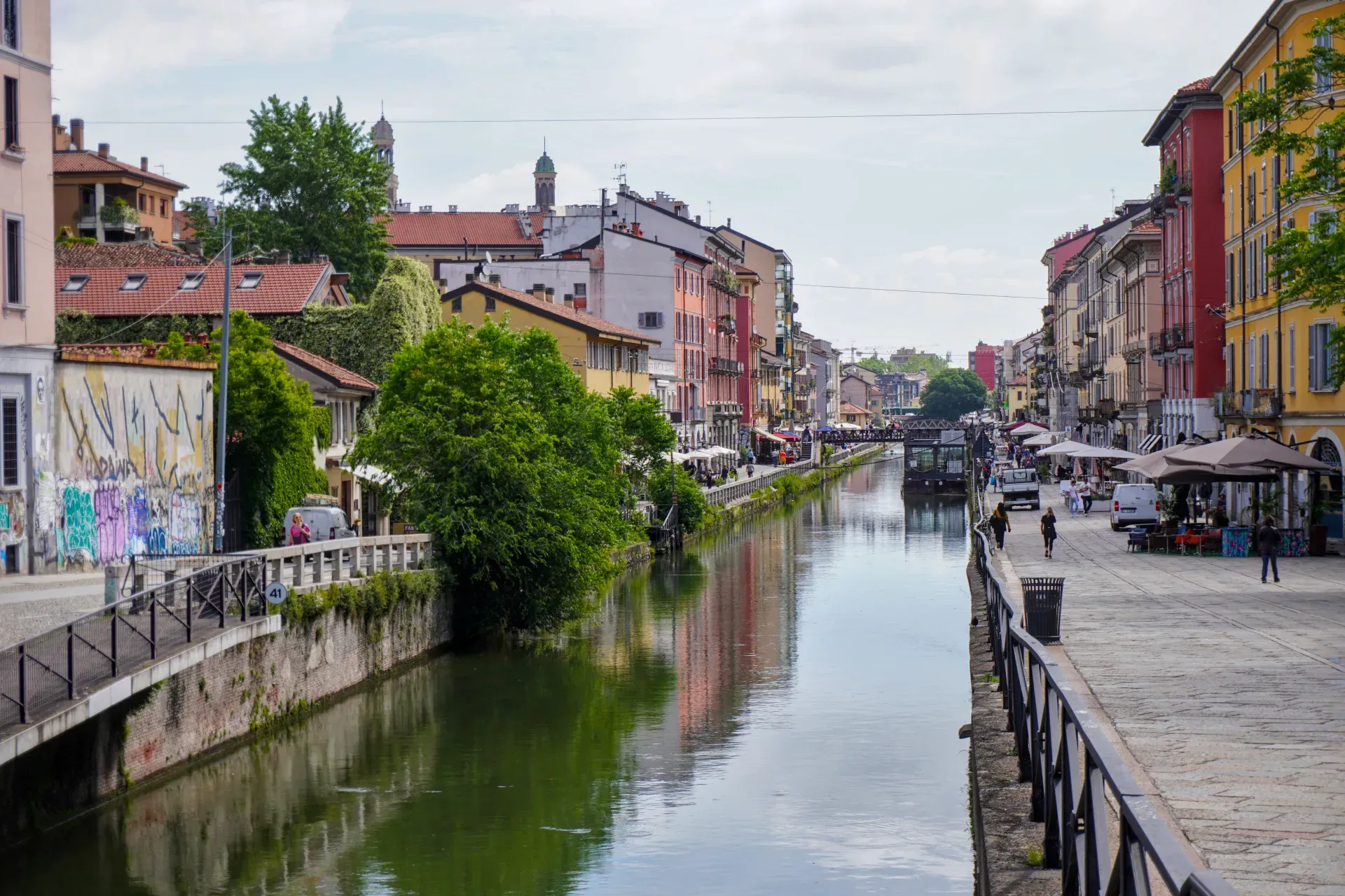 Ein Kanal im Mailänder Navigli-Viertel mit bunten Gebäuden, einer schwimmenden Struktur namens 'Square Garden', einem Boot und einem Roller.