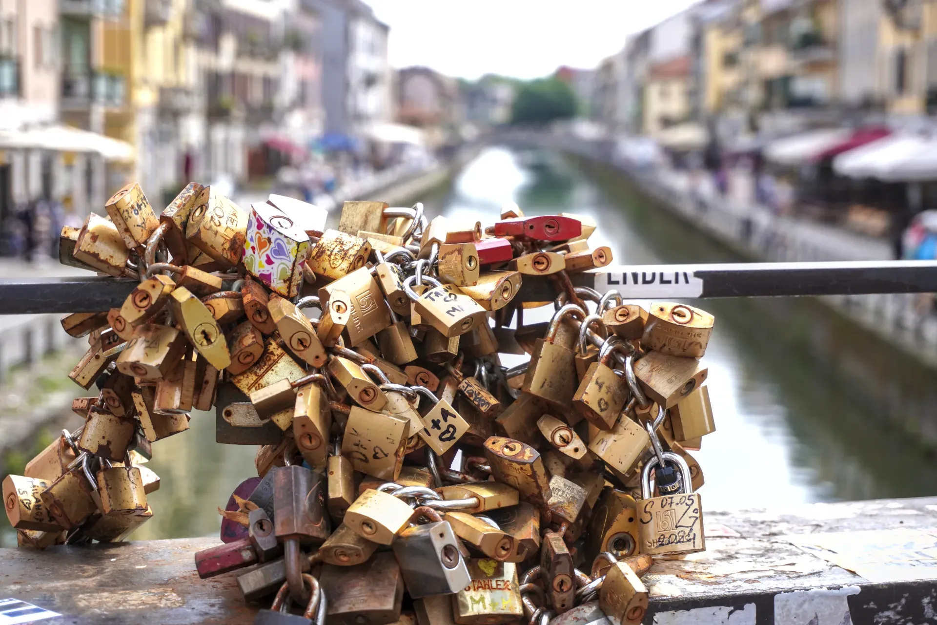 Une pile de cadenas patinés et colorés attachés à des chaînes sur la rambarde d'un pont, avec un canal et les bâtiments de Milan flous en arrière-plan.