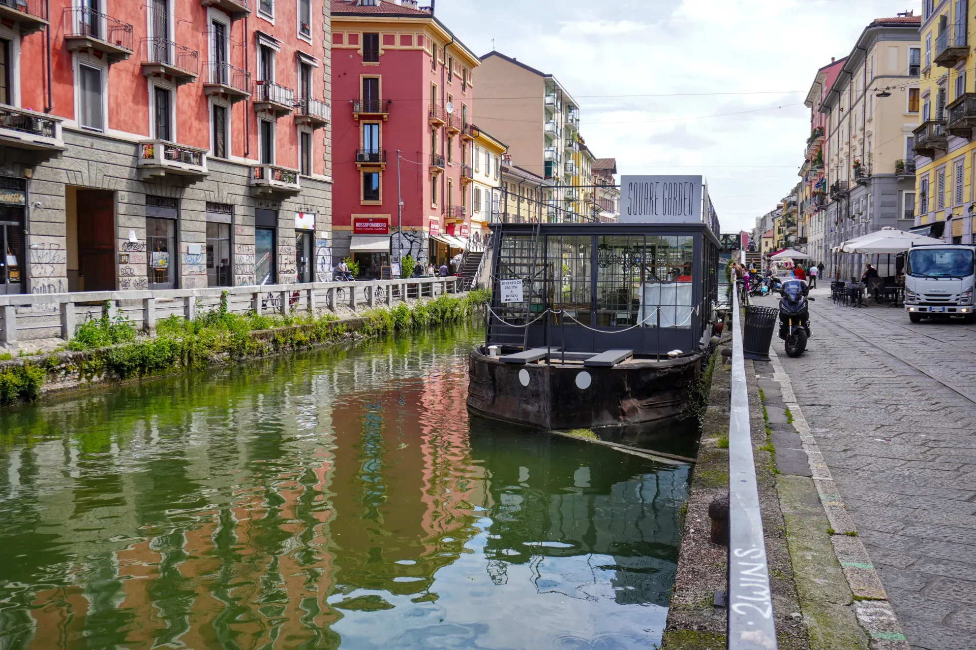 Un canale nel quartiere Navigli di Milano con edifici colorati su entrambi i lati, una passerella pedonale, persone che camminano, una barca sull'acqua e un ponte sullo sfondo.