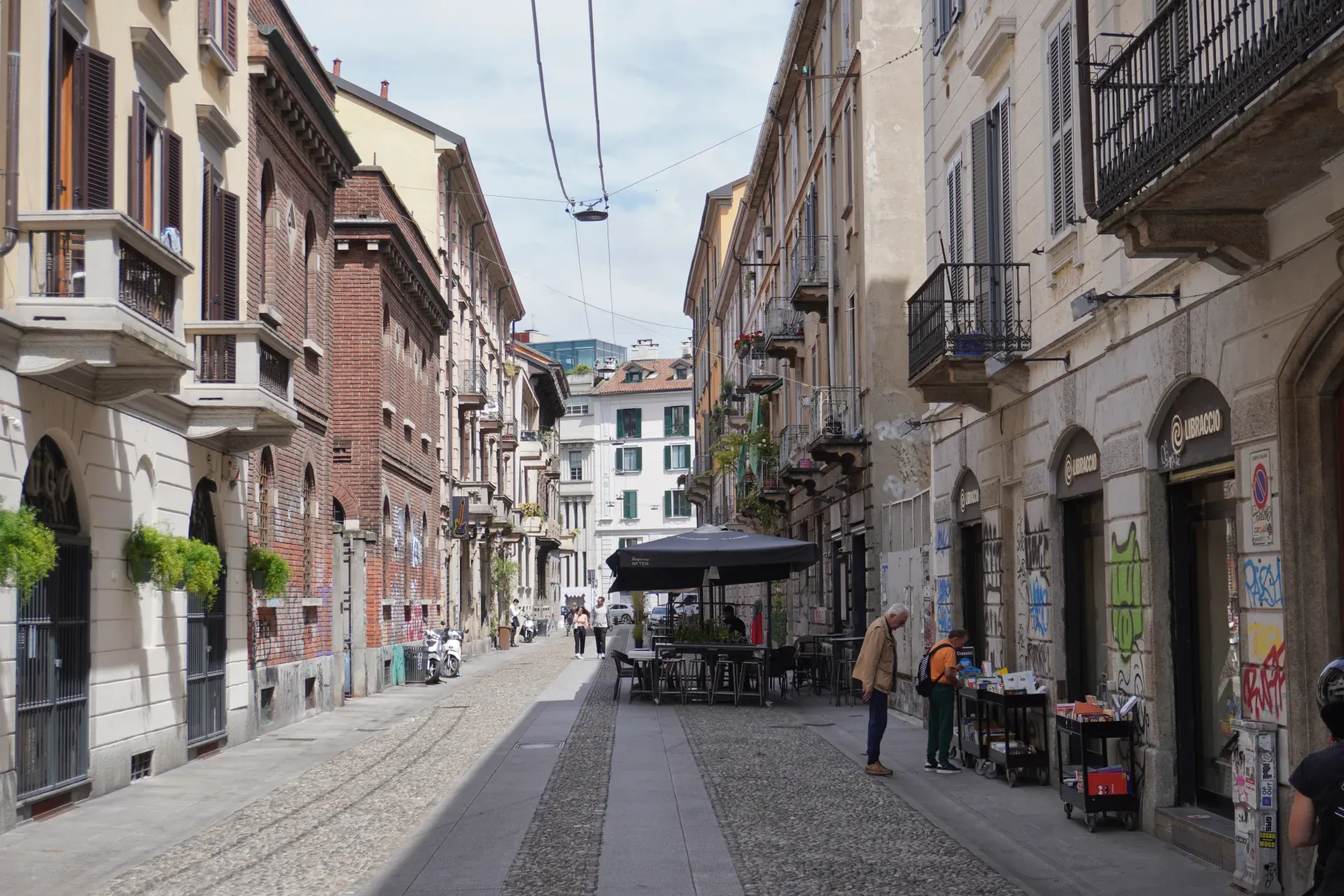 Un homme à vélo traverse une rue piétonne à Milan, Italie. Des bâtiments avec volets, balcons, graffitis et boutiques bordent la ruelle.