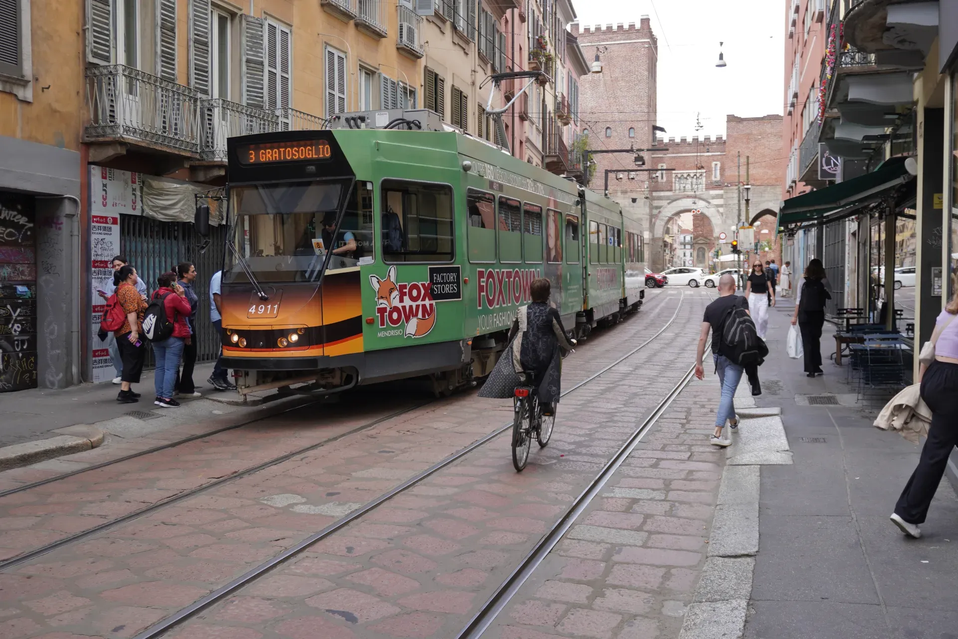 Un tram giallo su una strada acciottolata a Milano con edifici e caffè ai lati.