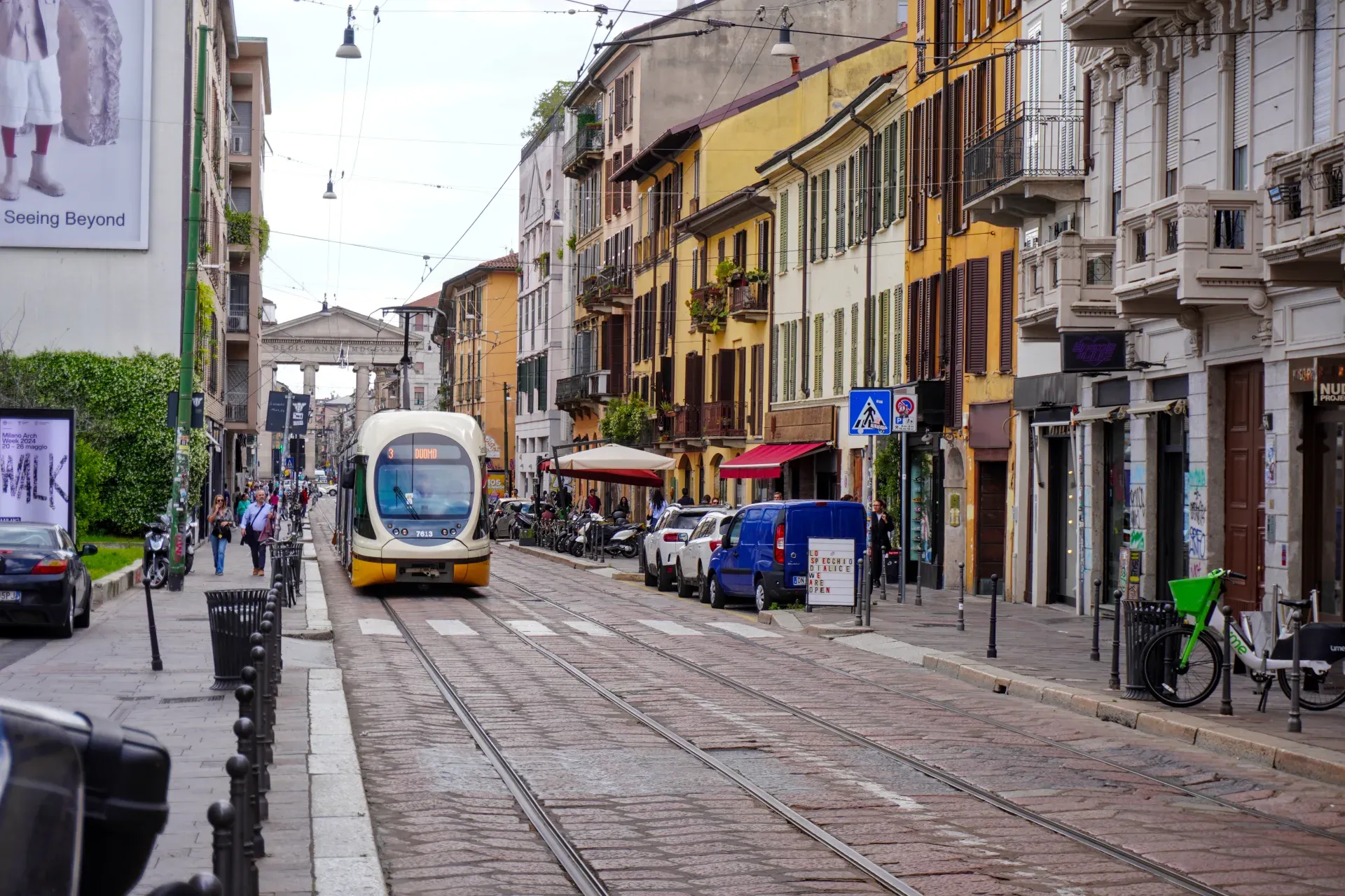 A yellow and white tram travels down a street in Milan with vintage buildings and shops lining the sides.