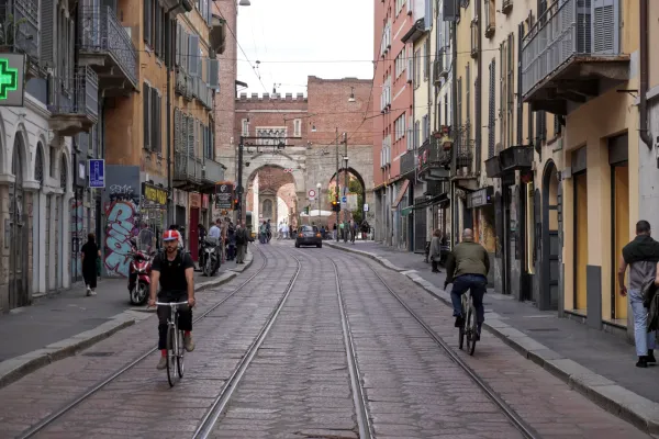 A green tram branded 'Foxtown' travels down a street in Milan, Italy, with pedestrians on the sidewalk and a cyclist nearby. Historic buildings and an archway are visible in the background.