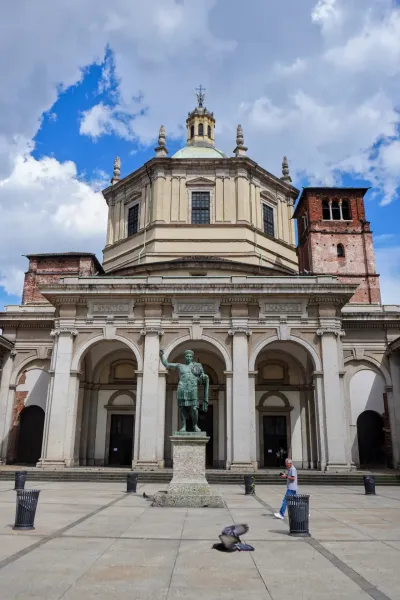 Statua cara Konstantina ispred Bazilike di San Lorenzo Maggiore sa kupolom i zvonarom pod oblacnim nebom.