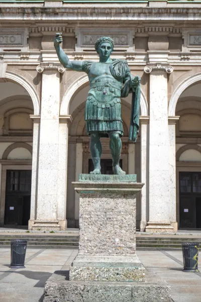 Eine hohe Bronzestatue von Kaiser Konstantin dem Großen in militärischer Rüstung, stehend auf einem Steinsockel vor einem Gebäude mit großen Bögen und klassischer Architektur in Mailand, Italien.
