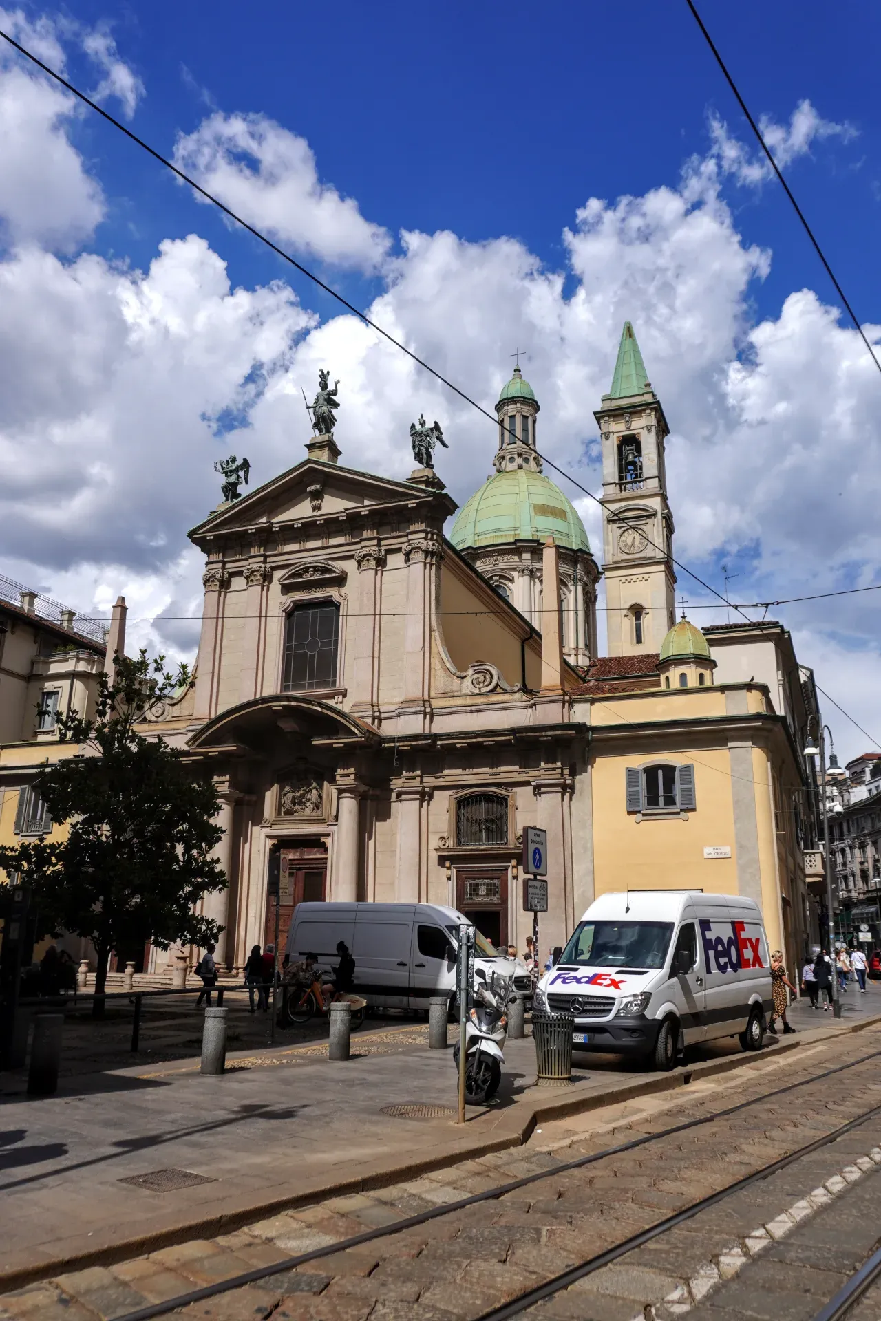 Une église européenne avec un dôme et une architecture classique se dresse dans une rue de Milan, avec des rails de tramway au premier plan et une camionnette de livraison FedEx garée sous un ciel bleu avec des nuages.