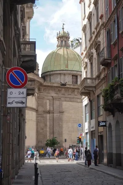 A European street in Milan, Italy, with a large green dome visible between buildings and people walking on a cobblestone street.