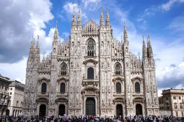 The ornate facade of Milan Cathedral, a large Gothic church, with many spires and statues, under a blue sky with clouds. A crowd of people is gathered in the square in front of the cathedral.