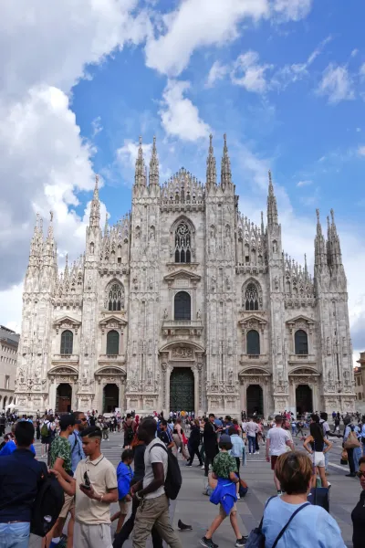 La façade ornée de la cathédrale de Milan, Italie, avec de nombreuses flèches et des détails en pierre sous un ciel bleu vif avec des nuages épars. Une foule de personnes est visible au premier plan sur la place.