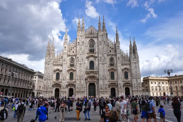 A wide shot of the Milan Cathedral, a grand Gothic cathedral with numerous spires and intricate stonework, against a cloudy sky. People are visible on the piazza in the foreground.