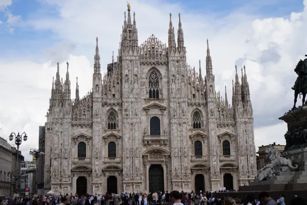 The intricate facade of Milan Cathedral (Duomo di Milano) under a cloudy sky, with a bronze equestrian statue and trees in front, and a crowd in the square.