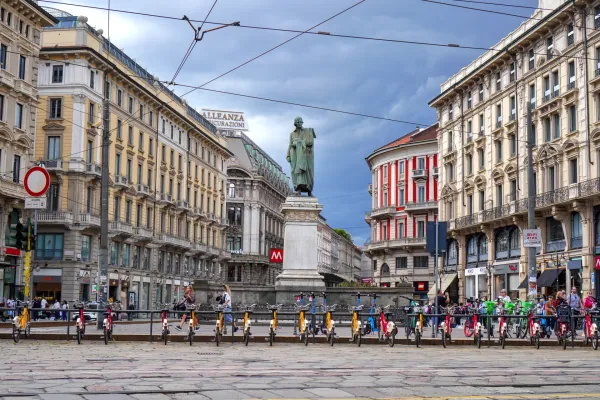 A street in Milan, Italy, with ornate buildings lining the sides, a bronze statue on a pedestal in the center, and numerous yellow and red electric scooters and bicycles parked in a row.