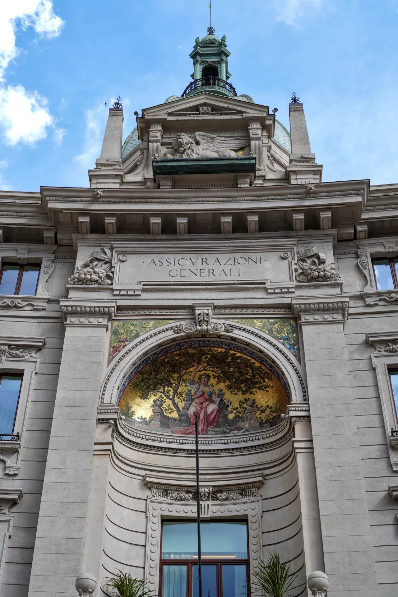Una vista detallada de la fachada superior del edificio Assicurazioni Generali en Milán, que muestra elementos arquitectónicos clásicos, un mosaico y un león alado.