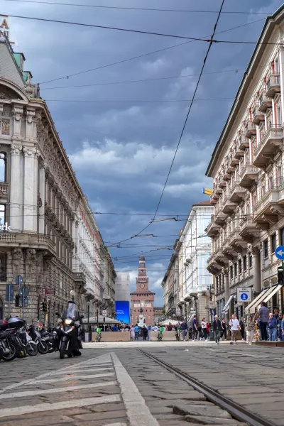 Eine Straße in Mailand, Italien, mit einer Straßenbahn, die auf Kopfsteinpflasterschienen fährt, flankiert von historischen Gebäuden. Das Castello Sforzesco ist im Hintergrund unter einem bewölkten Himmel sichtbar.