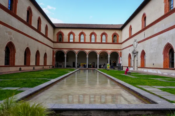 Inner courtyard view of the Castello Sforzesco in Milan, Italy, featuring arcade-lined buildings, manicured lawns, and a reflective water feature.