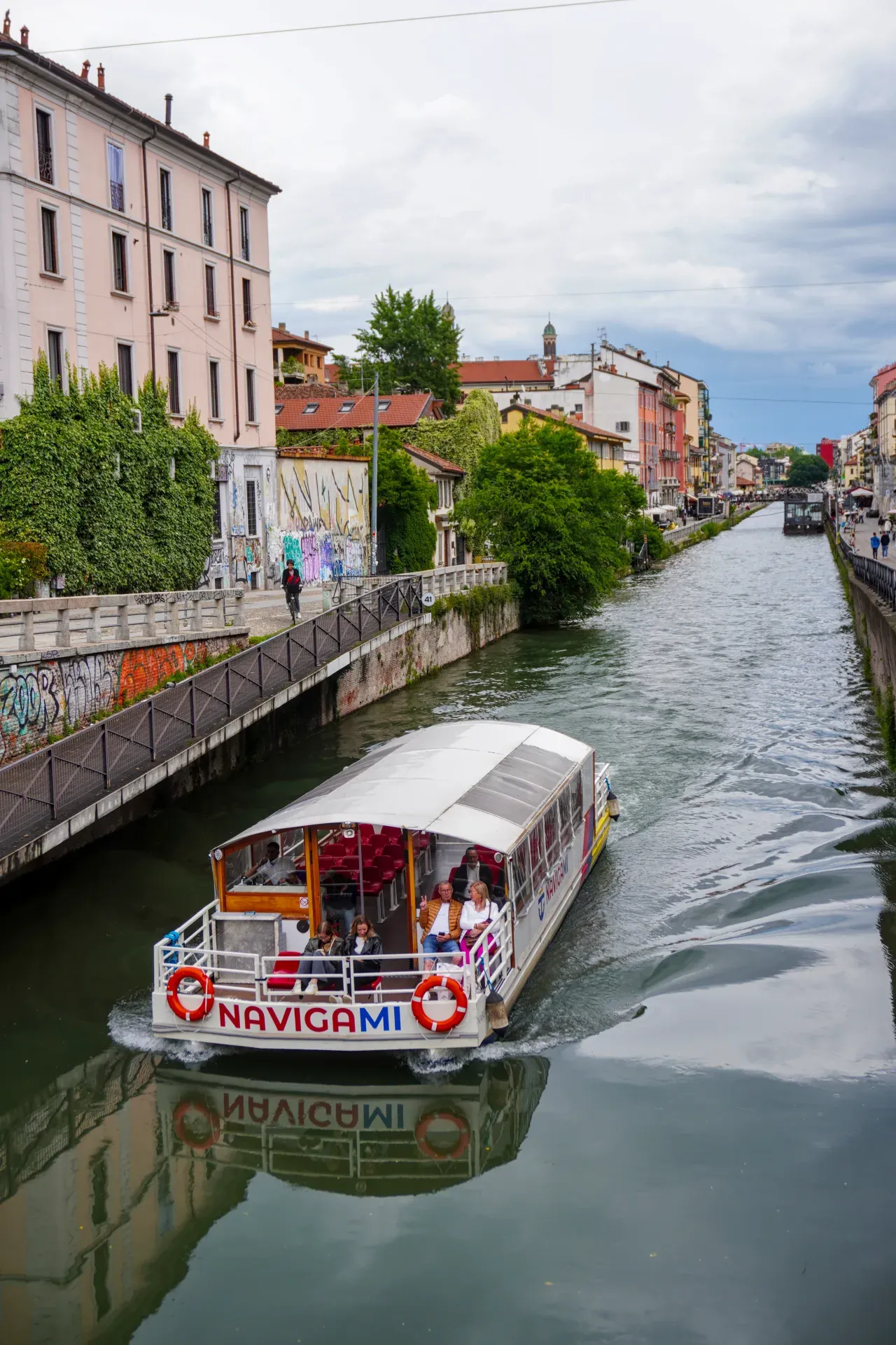 A tour boat named "Navigami" sails on a canal in Milan, Italy, with passengers on board. Buildings and green spaces line the canal, and the reflection of the boat is visible in the water.