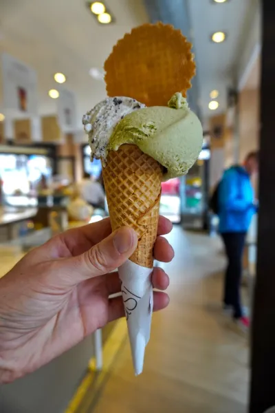 Eine Hand hält einen Waffelhorn mit drei Kugeln Gelato, darunter eine Pistazie und eine Keks-Creme, garniert mit einer runden Waffel. Im verschwommenen Hintergrund sind Menschen in einer Gelateria in Mailand zu sehen.