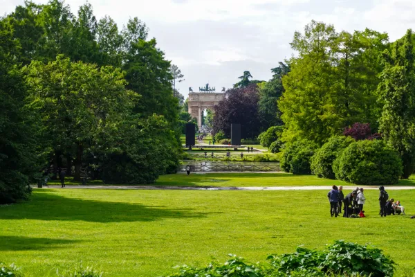 Der Arco della Pace, ein großer Triumphbogen, steht am Ende einer langen Allee im Parco Sempione. Vor dem Bogen sind Menschen zu sehen, die auf Rasenflächen und Wegen spazieren gehen, während grüne Bäume die Szene säumen.