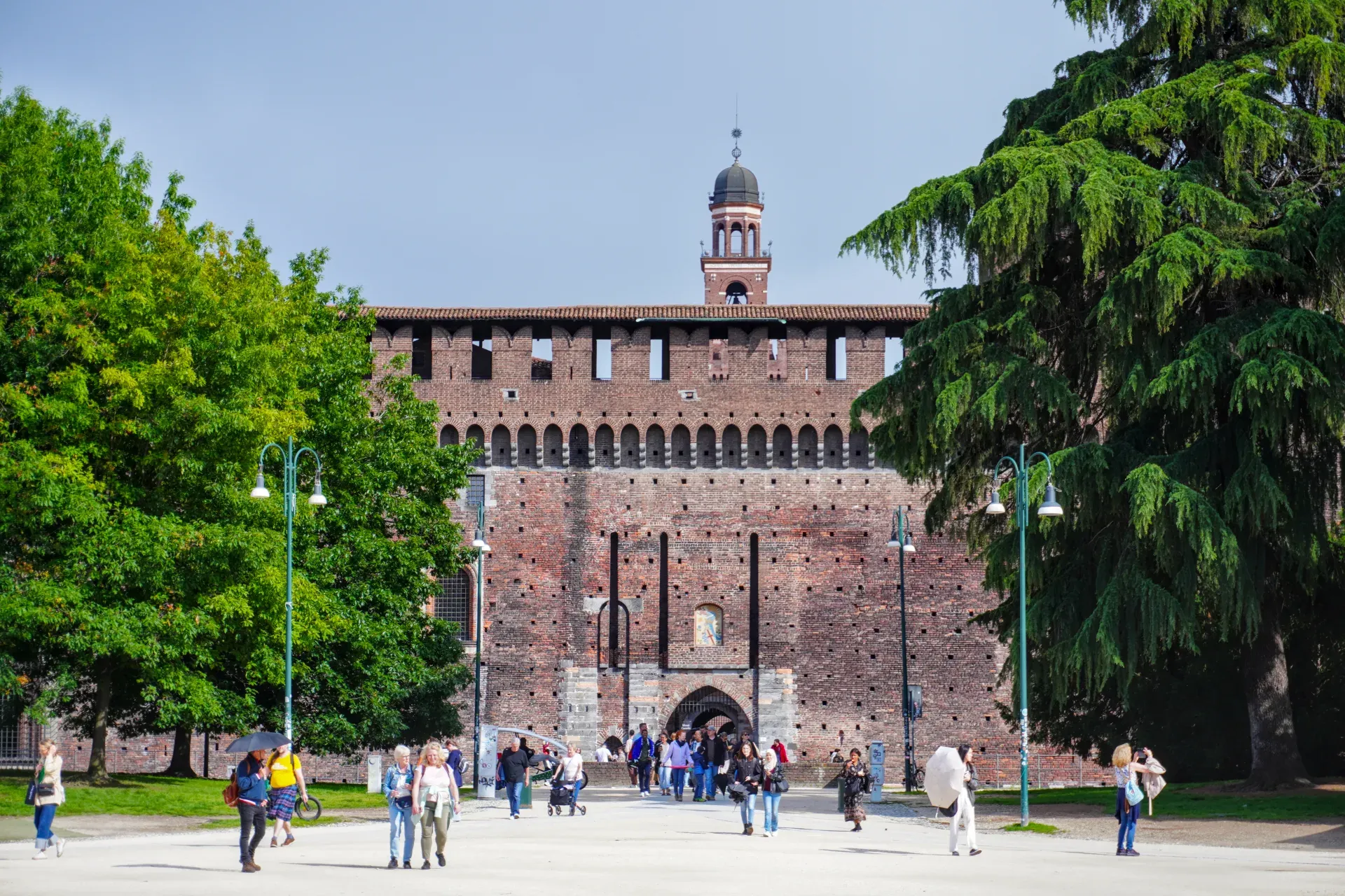 Des gens se promènent devant le Château Sforza à Milan, Italie, avec de grands arbres encadrant la vue.