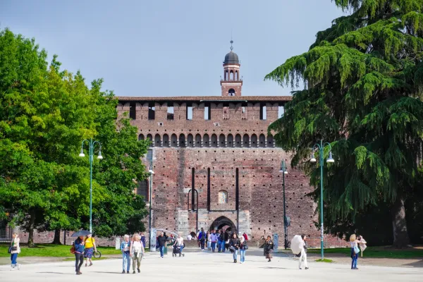 Ljudi šetaju ispred zamka Sforca u Milanu, Italija, sa velikim drvećem koje uokviruje pogled.