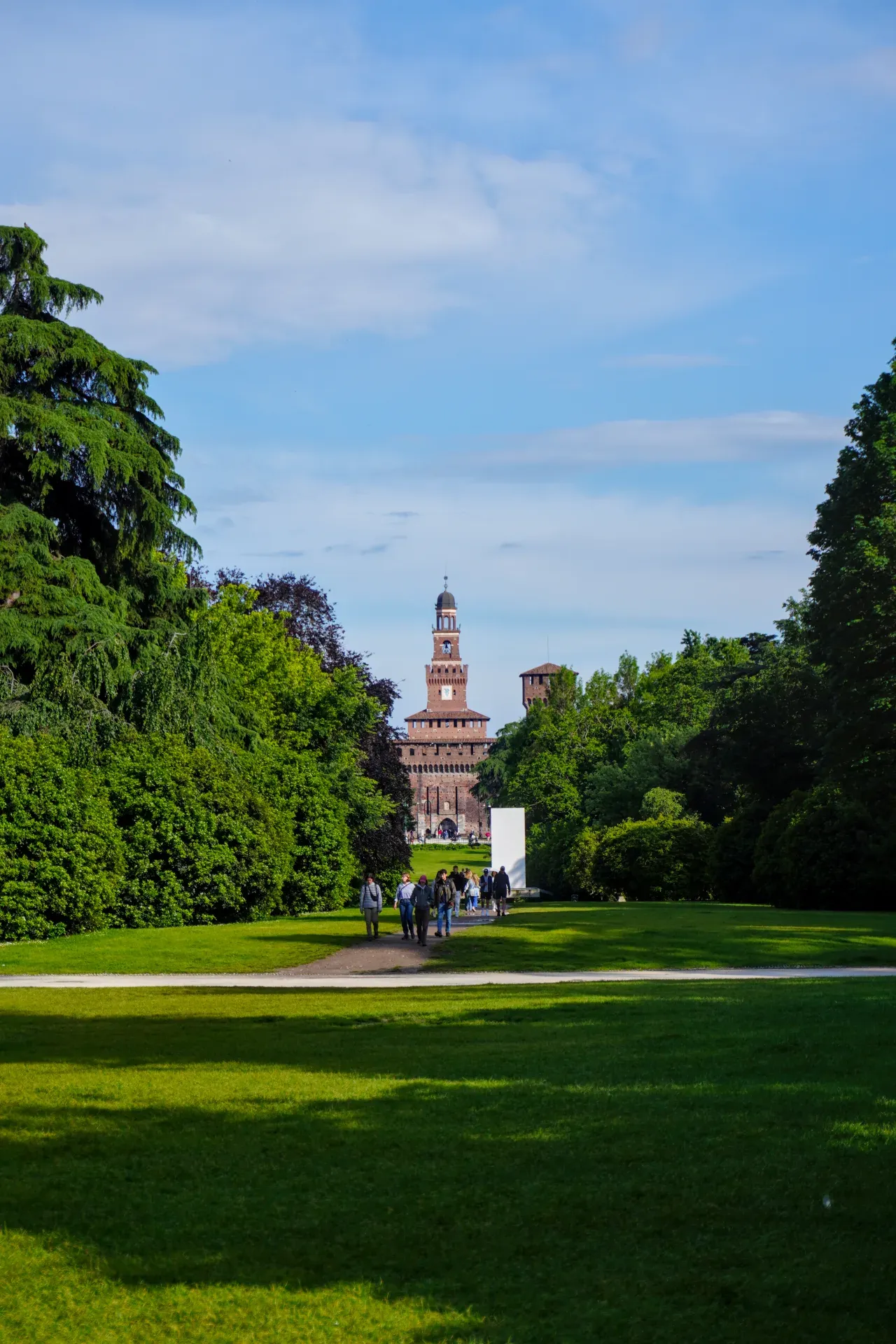 Ein Weg durch einen üppig grünen Park in Mailand führt zum imposanten Schloss Sforzesco mit seinem markanten Uhrturm.