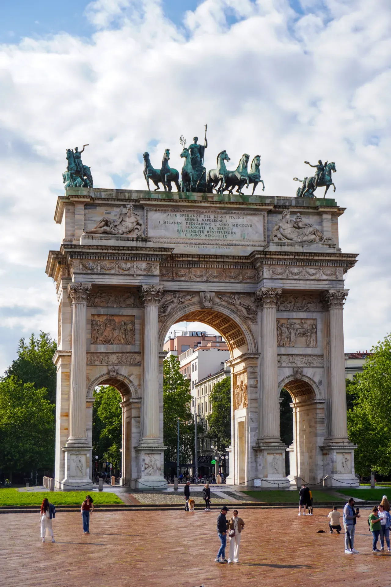 L'imposant Arco della Pace à Milan, Italie, sous un ciel nuageux, avec des gens au premier plan et des arbres entourant le monument.