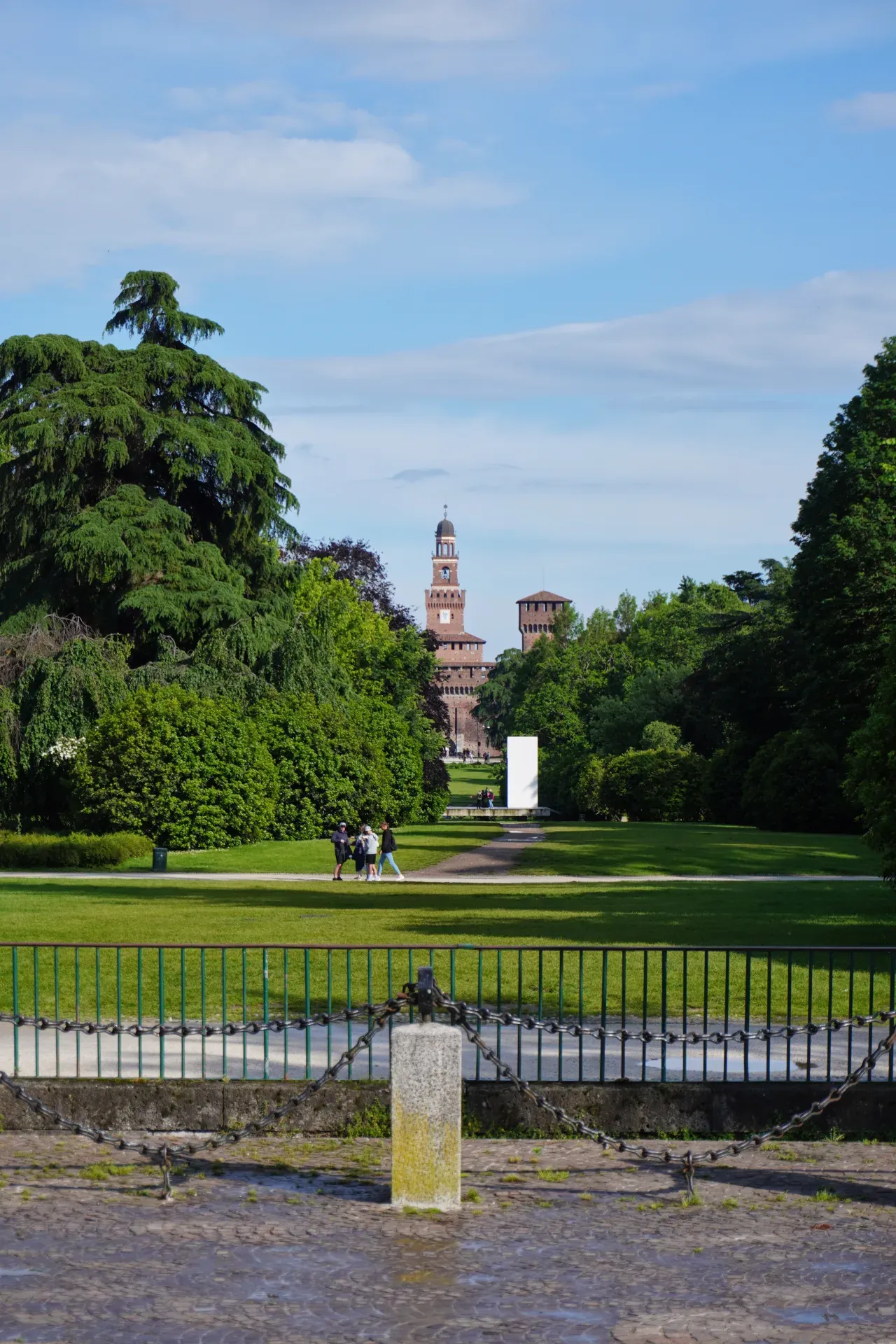 La torre del Castello Sforzesco si erge sopra gli alberi nel Parco Sempione, Milano, con persone che camminano su un sentiero in primo piano.