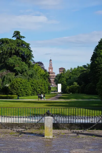 Turm des Schlosses Sforza ragt über Bäumen im Parco Sempione, Mailand, mit Menschen, die im Vordergrund auf einem Weg spazieren.