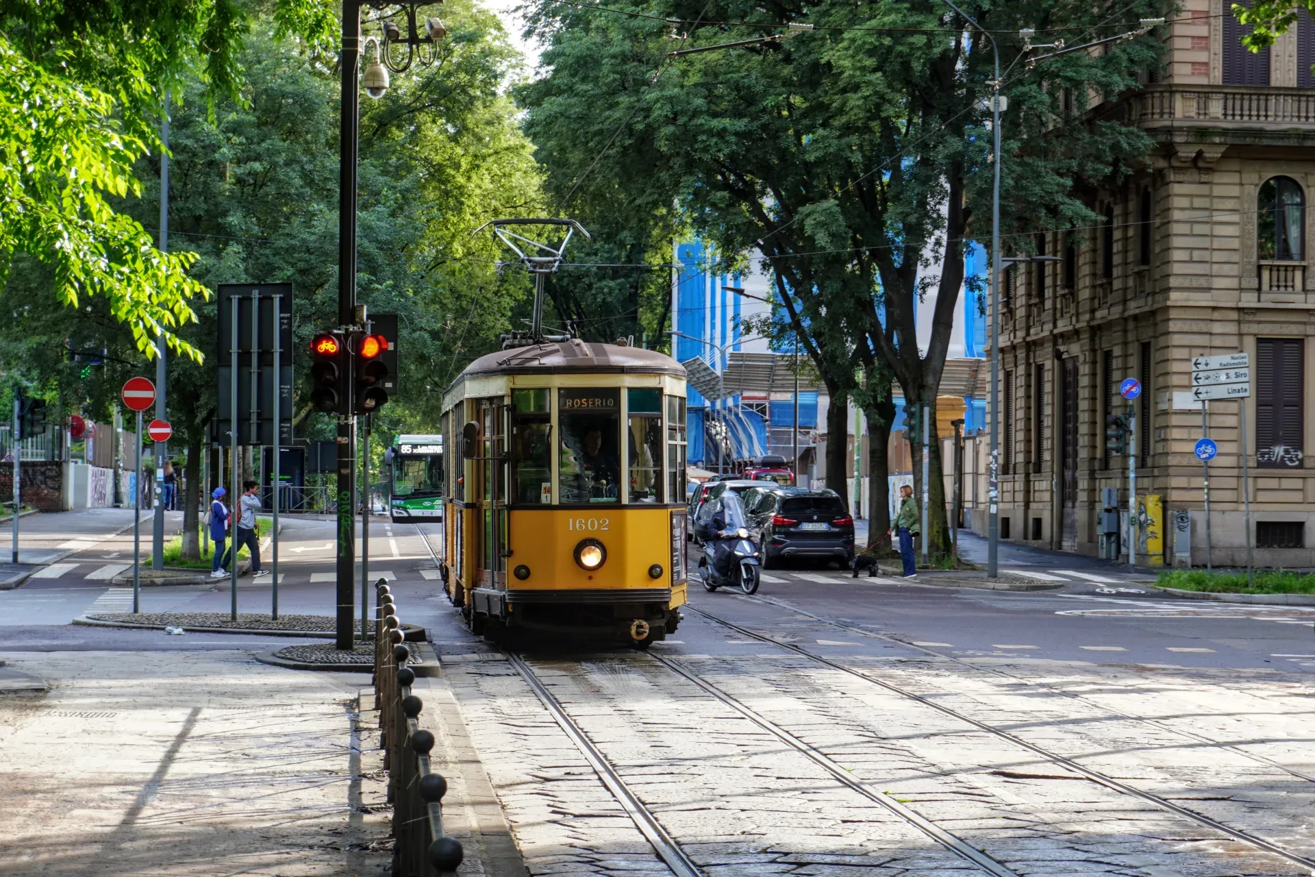 Un tram giallo vintage, numero 1602, viaggia sui binari lungo una strada di Milano, con alberi, edifici, un autobus, un'auto e uno scooter sullo sfondo.