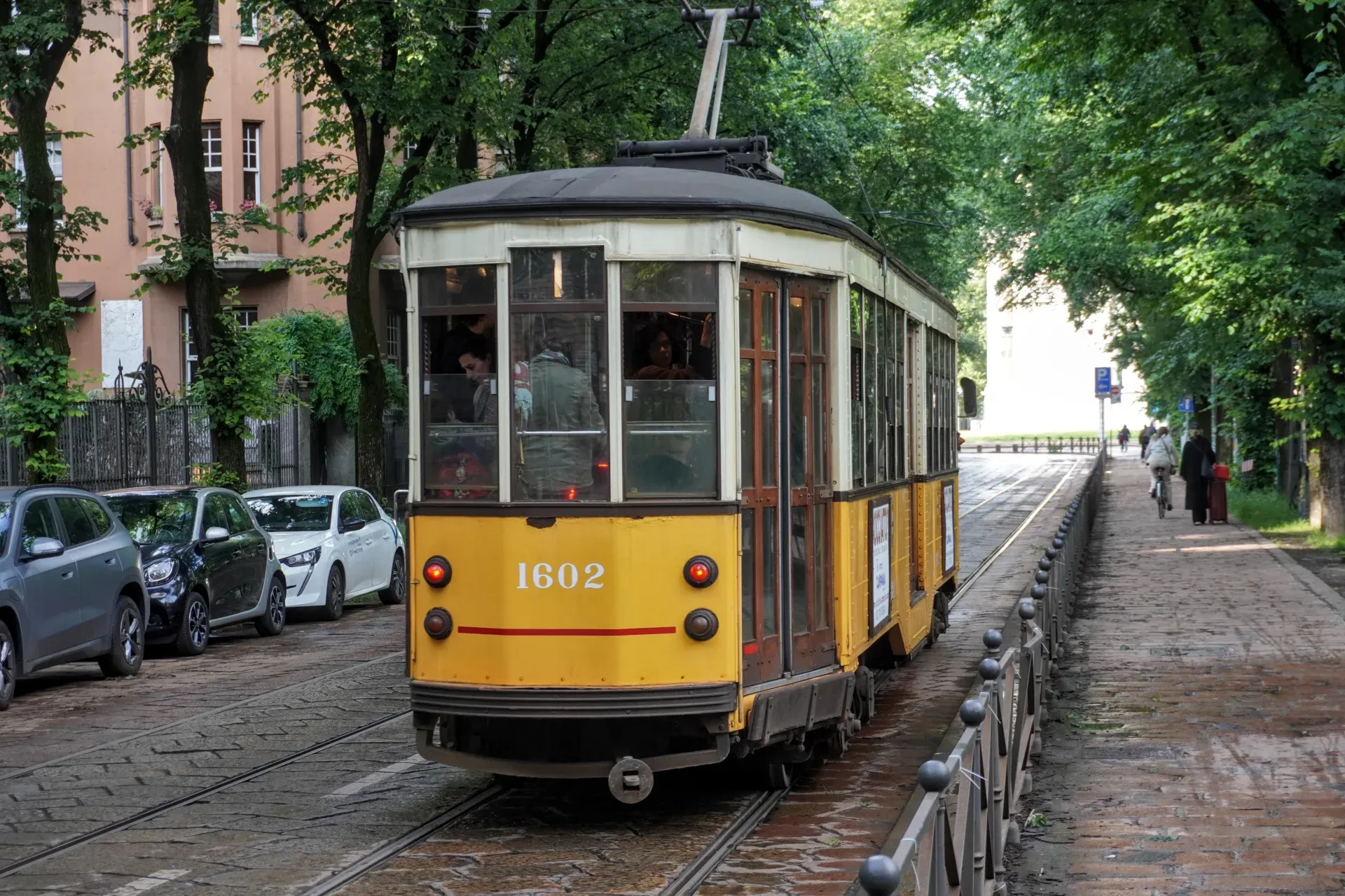 Un tramway jaune et beige circule dans une rue de Milan, Italie, avec des bâtiments historiques ornés d'un côté et un restaurant moderne en verre de l'autre. Des vélos sont garés sur le trottoir.