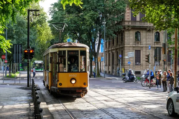 Eine gelbe Vintage-Straßenbahn, Nummer 1602, bewegt sich auf Gleisen auf einer Straße in Mailand. Auf der Vorderseite der Straßenbahn steht 'ROSERIO' angezeigt. Im Hintergrund sind Gebäude, Bäume, Ampeln und Menschen zu sehen.