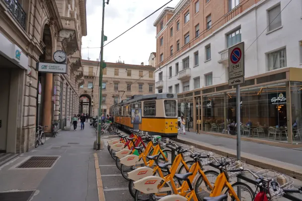 A street in Milan, Italy, with a yellow tram on tracks, a row of orange rental bikes, pedestrians on the sidewalk, and buildings with shops and cafes.