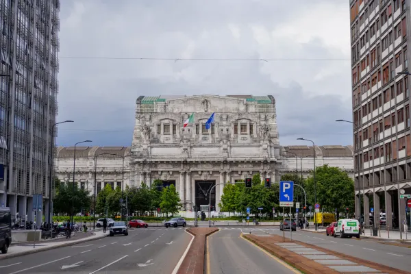 The impressive facade of Milan Central Station with Italian and European flags, surrounded by trees and with traffic on the street in the foreground.