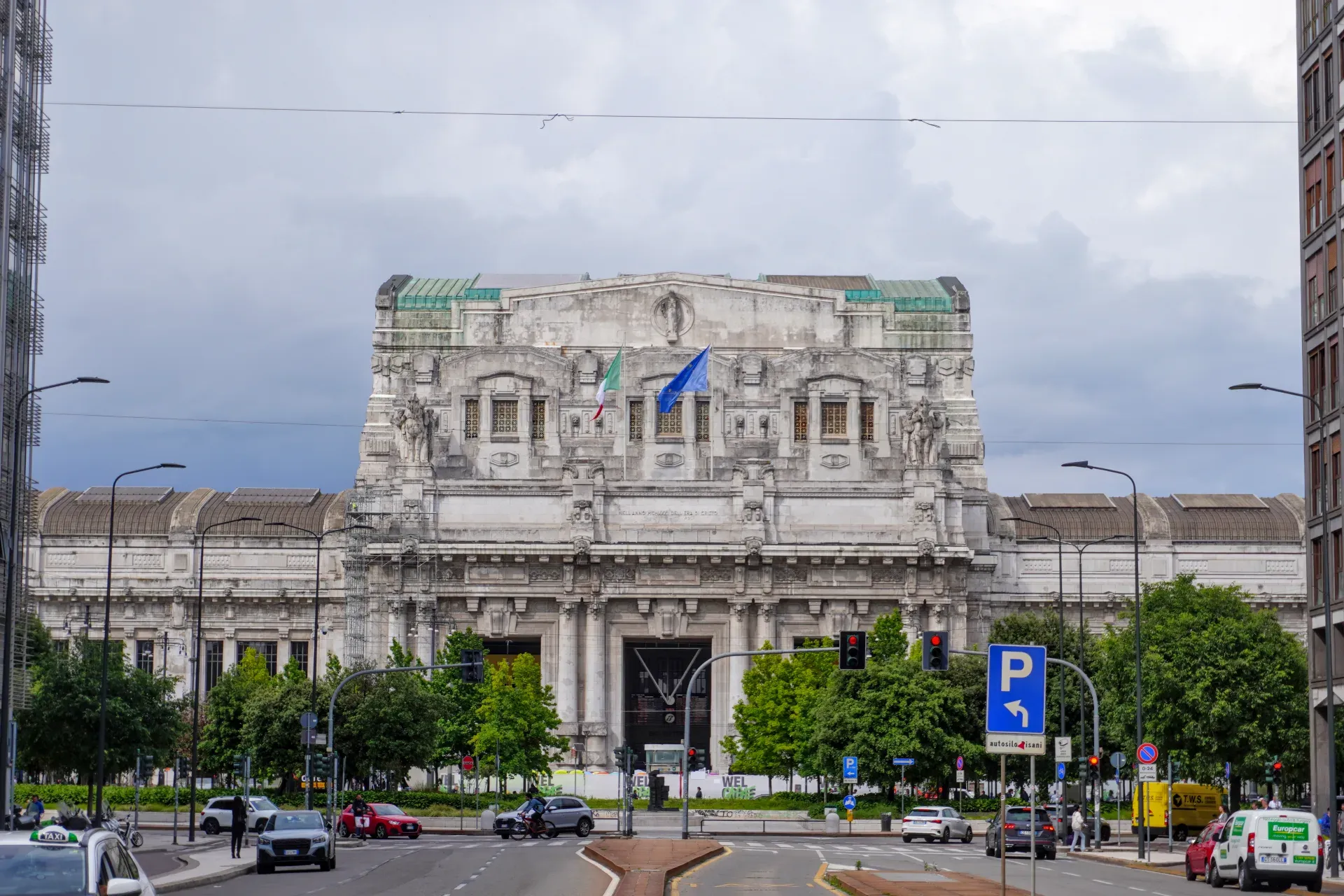 Impresivna fasada Železničke stanice Milano Centrale sa italijanskim i zastavama EU tokom oblačnog dana, sa automobilima i drvećem na ulici u prvom planu.