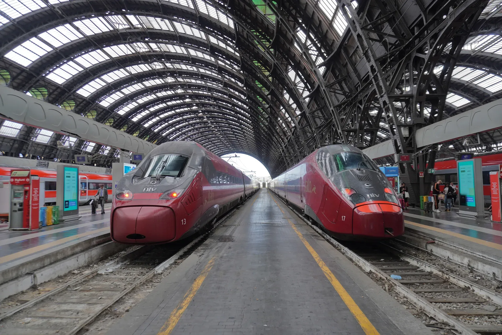 Two modern red high-speed trains wait on the platforms at Milan Central Station under its grand arched glass roof.