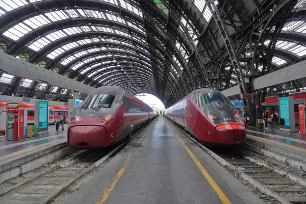 Two modern red high-speed trains wait on the platforms at Milan Central Station under its grand arched glass roof.