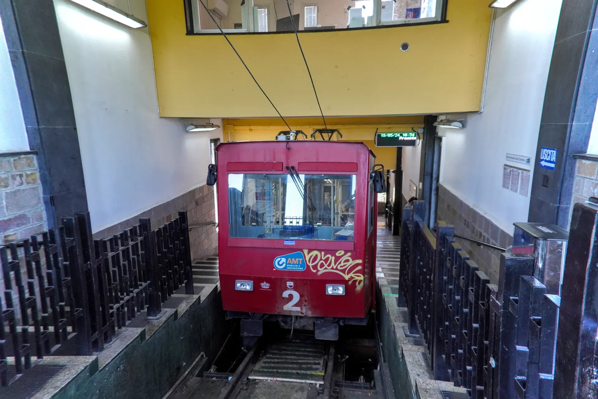 Un train de funiculaire rouge est garé dans une gare avec des escaliers menant à la sortie et des portillons de billetterie au premier plan.