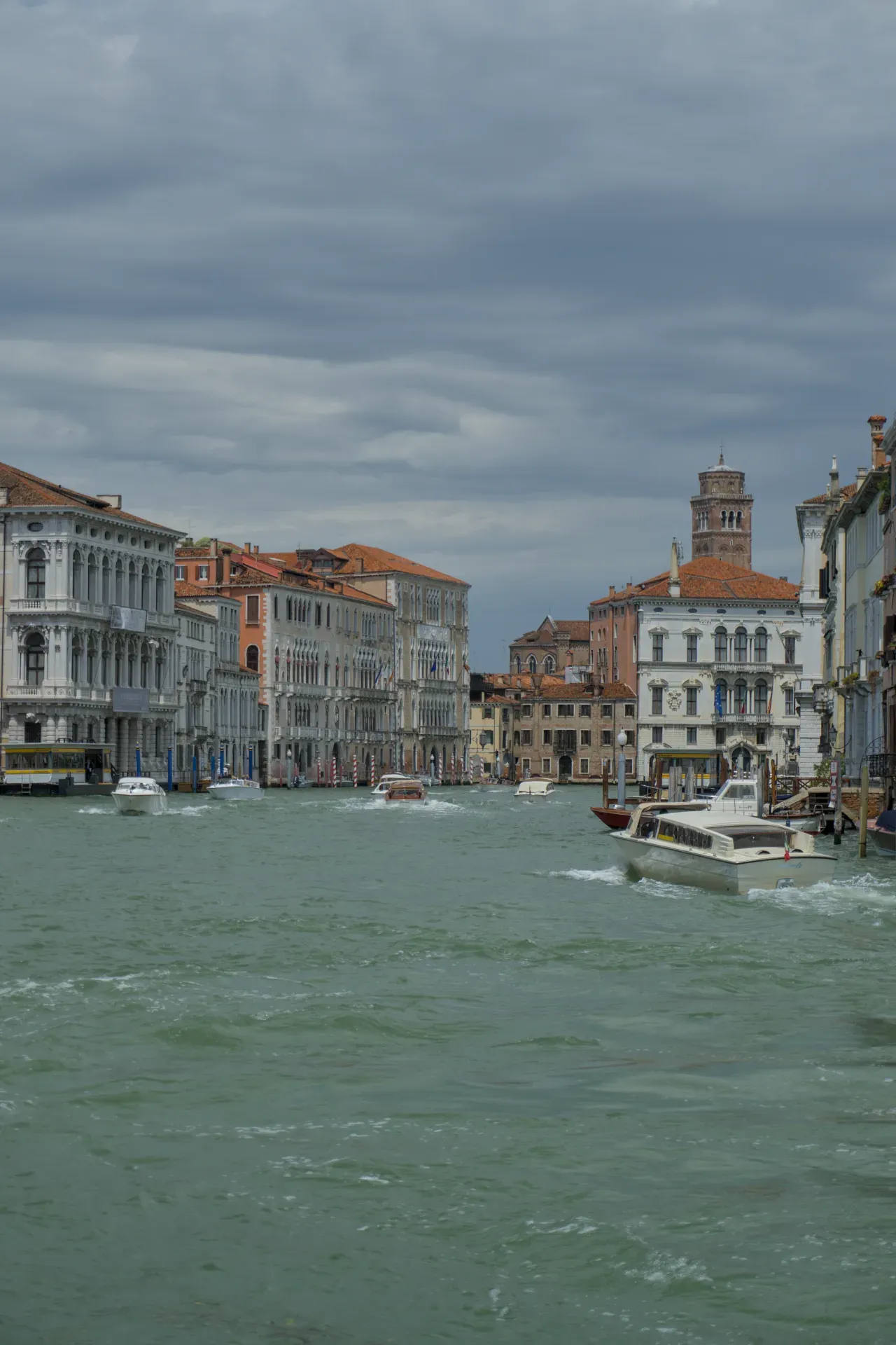 Barcos en el Gran Canal de Venecia, Italia, con edificios ornamentados y un campanario al fondo bajo un cielo nublado.