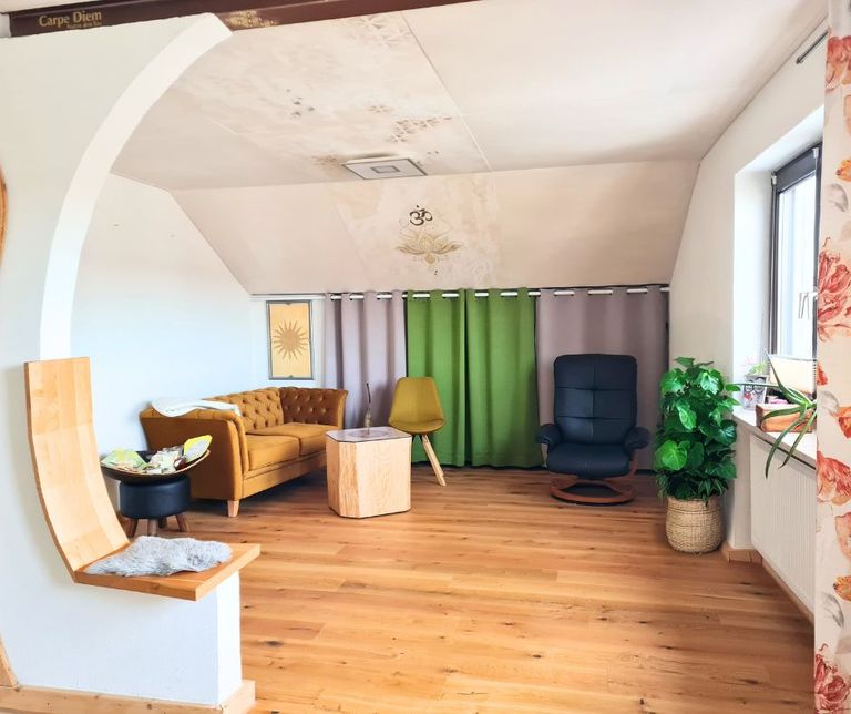 Kleiner Seminarraum mit einem braunen Sofa, einem gelben Stuhl, einem schwarzen Sessel, einem kleinen Holztisch und einem grünen Vorhang vor einem Fenster, mit Holzboden und weißen Wänden.
