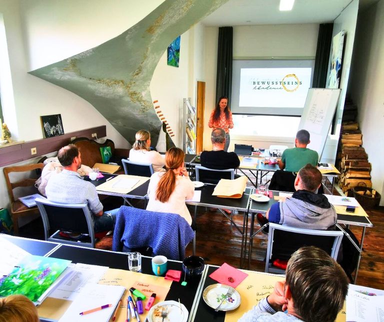 Mehrere Personen sitzen an Tischen in einem Seminarraum und hören einer Frau zu, die vorne vor einer Leinwand mit Präsentation steht.