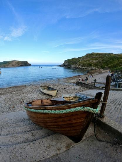 A serene beach scene featuring a wooden boat moored on a pebbly shore, with several smaller boats nearby. The calm water reflects a clear blue sky, while gentle hills frame the background. A few people can be seen enjoying the beach area.