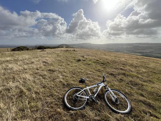 A white mountain bike resting on grassy terrain with a panoramic view of distant hills and clouds in a blue sky. The sun is shining brightly, casting light across the landscape.