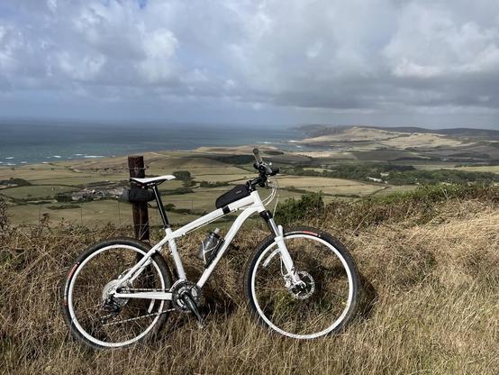 A white mountain bike is parked on a hillside overlooking a coastal landscape. The view features rolling green fields, a distant beach, and the ocean under a partly cloudy sky.