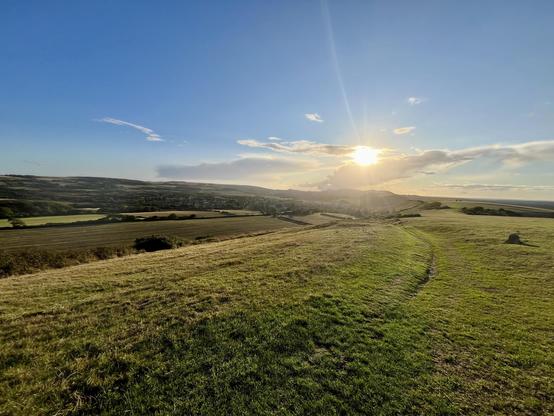 A scenic view of rolling hills and fields under a bright blue sky. The sun is setting in the background, casting warm light across the landscape. A winding path leads through the grassy terrain.