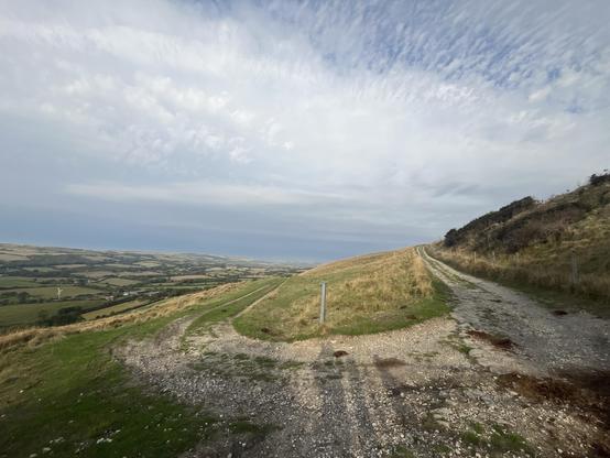 A scenic landscape featuring two dirt paths winding through green hills, with patches of grass and sparse shrubs. The background shows rolling valleys leading to the sea, under a partly cloudy sky.
